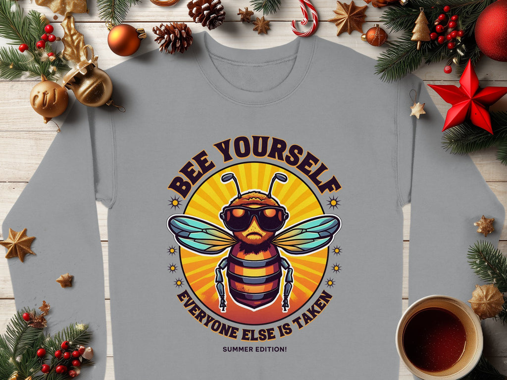 A Garment Graphics gray unisex T-shirt shows a cartoon bee in sunglasses and a crown with Bee Yourself, Everyone Else is Taken, Summer Edition text. It’s displayed on a wooden surface with festive decorations and a coffee cup.