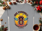 A Garment Graphics gray unisex T-shirt shows a cartoon bee in sunglasses and a crown with Bee Yourself, Everyone Else is Taken, Summer Edition text. It’s displayed on a wooden surface with festive decorations and a coffee cup.