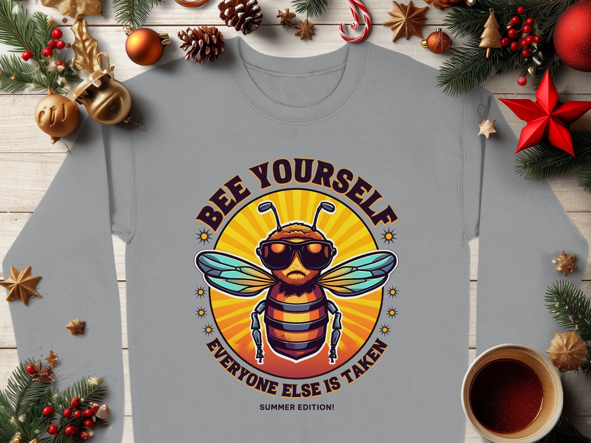 A Garment Graphics gray unisex T-shirt shows a cartoon bee in sunglasses and a crown with Bee Yourself, Everyone Else is Taken, Summer Edition text. It’s displayed on a wooden surface with festive decorations and a coffee cup.