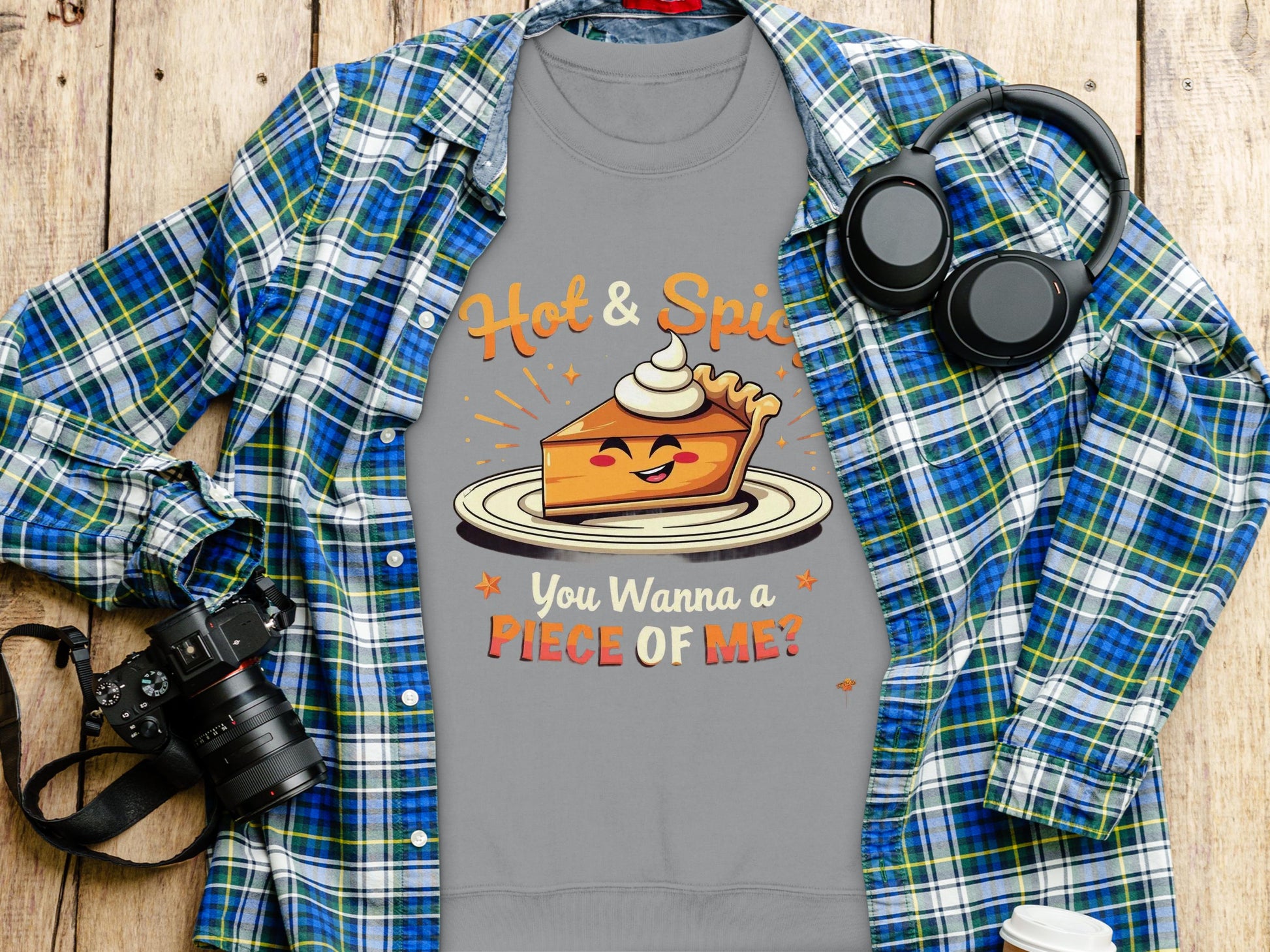 A gray Garment Graphics pie graphic sweatshirt showcases a smiling cartoon pie slice with whipped cream and bold text, Hot & Spicy. You Wanna a Piece of Me? It sits on a plaid shirt amid a camera and headphones on the wooden surface.