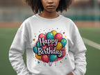 A young person models a vibrant Happy Birthday sweatshirt by Garment Graphics, featuring colorful balloons and a cheerful message. The design radiates celebration and joy against a blurred outdoor backdrop, offering unisex sizing for everyone.