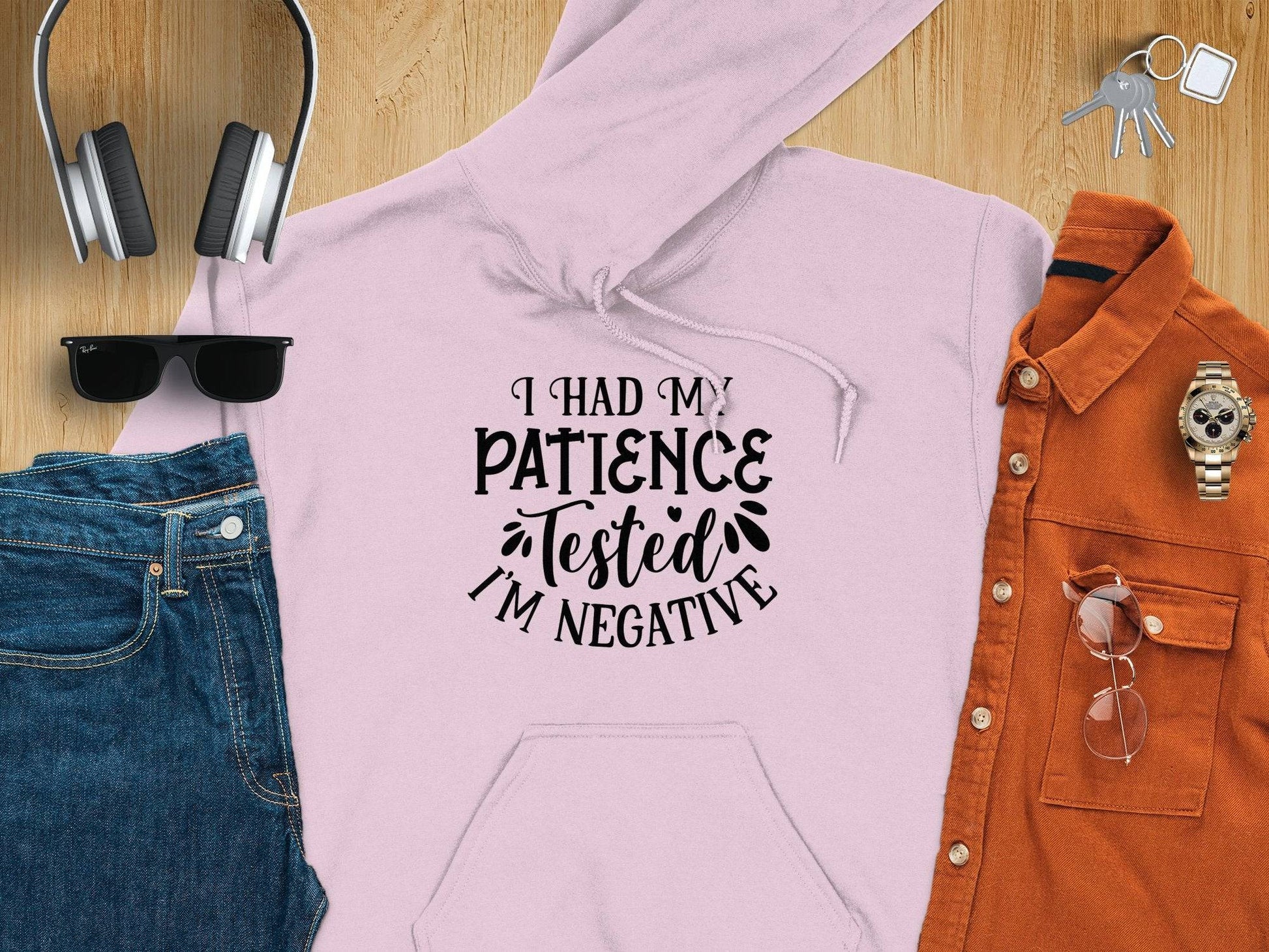 A light pink Patience Tested Hoodie by Garment Graphics, featuring I Had My Patience Tested, Im Negative, rests on wood. The medium-heavy fabric offers warmth, accompanied by headphones, black sunglasses, jeans, an orange shirt, watch, and keys.