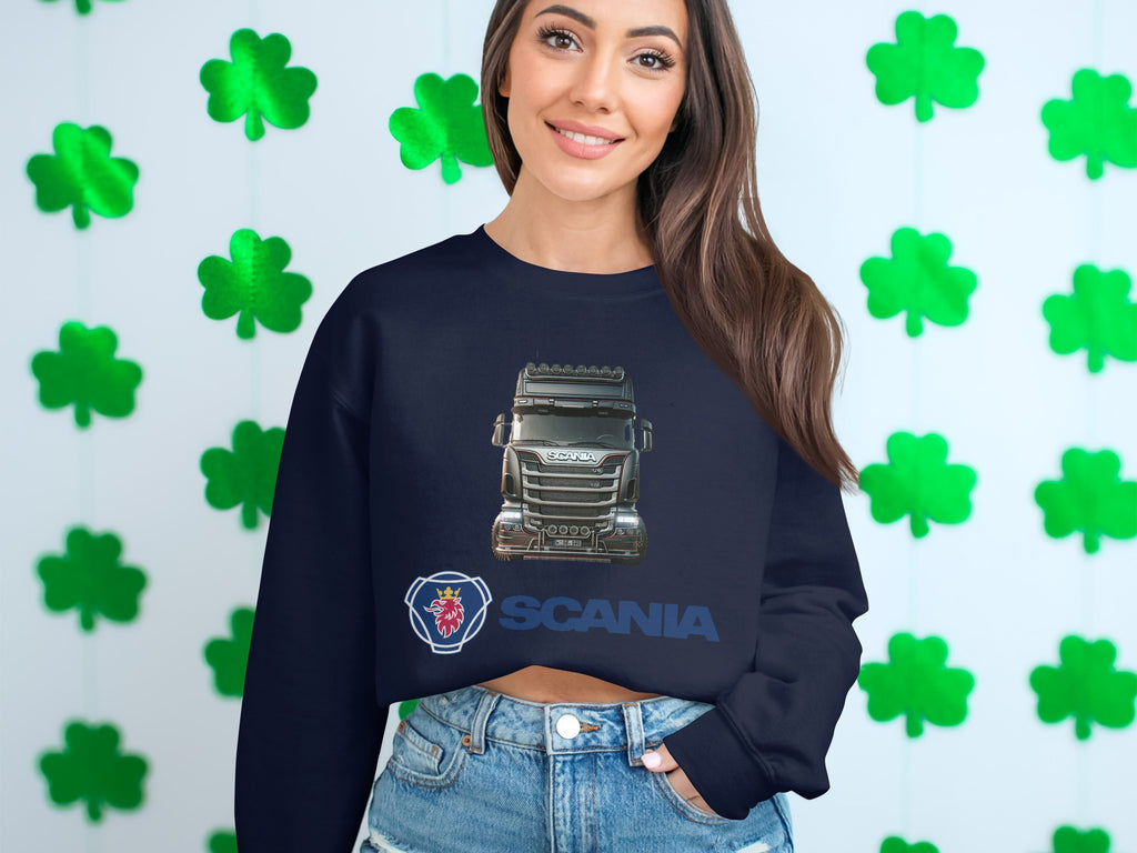 In front of a shamrock-covered wall, a person with long hair smiles in a Garment Graphics crewneck sweatshirt featuring SCANIA and a truck. Their casual stance, with one hand in their jean pocket, reflects the carefree spirit of truck enthusiasts.