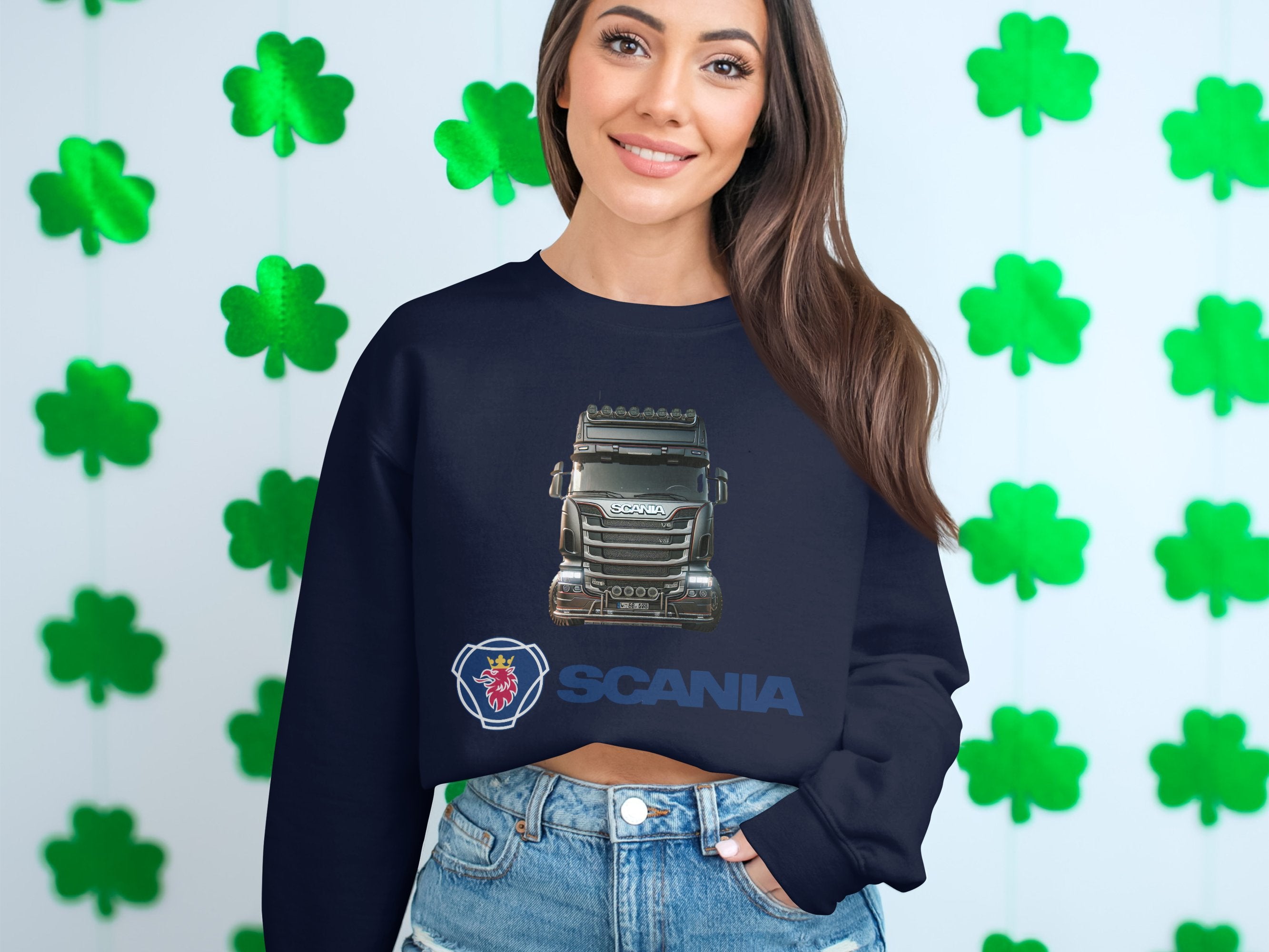 In front of a shamrock-covered wall, a person with long hair smiles in a Garment Graphics crewneck sweatshirt featuring SCANIA and a truck. Their casual stance, with one hand in their jean pocket, reflects the carefree spirit of truck enthusiasts.