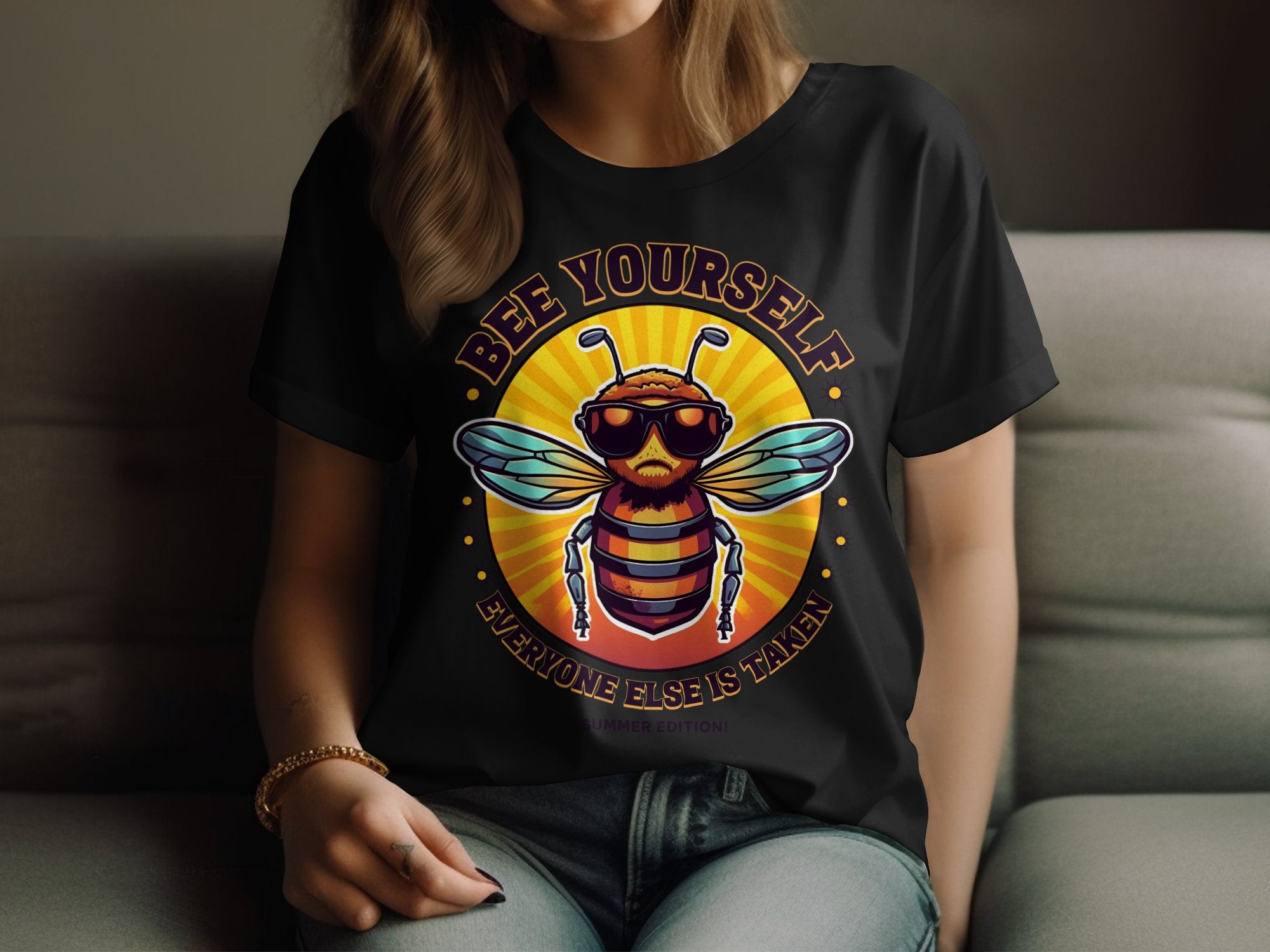 A person is sitting, wearing a black T-shirt from Garment Graphics Bee Yourself Summer Edition. It showcases a bee with sunglasses and the message, Bee Yourself, Everyone Else Is Taken, in womens sizing as they rest their hand on their knee.