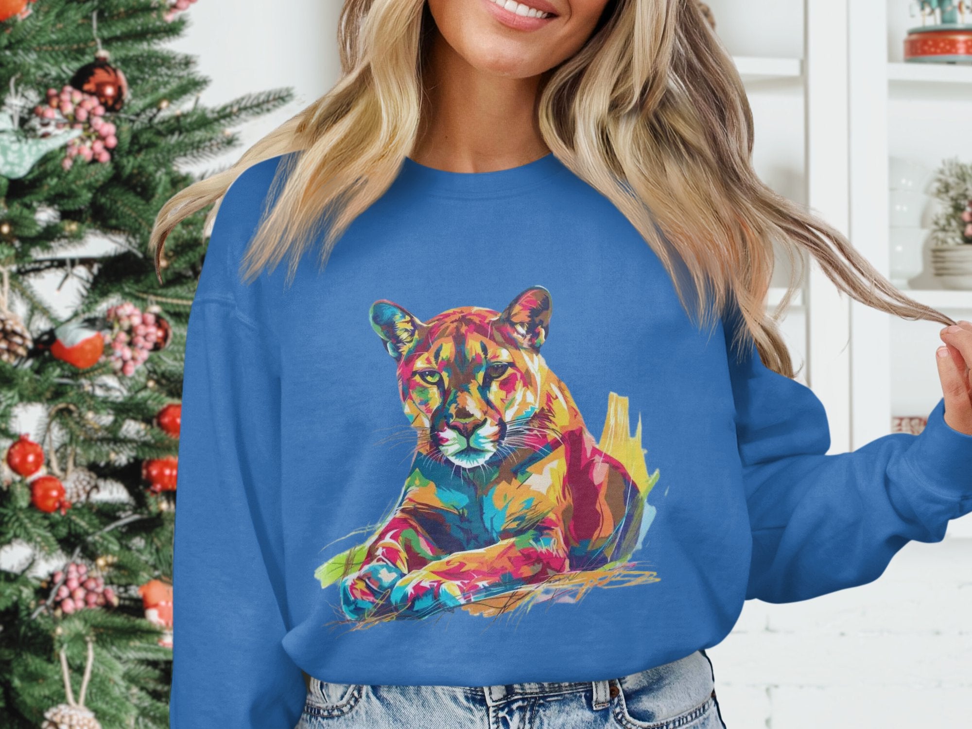 A woman with long, light hair stands by a decorated Christmas tree, wearing Garment Graphics blue unisex sweatshirt featuring a vibrant big cat design. She holds a hair strand with her left hand, with white shelves in the background.