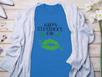 The Garment Graphics blue graphic T-shirt features green lips and clovers with Happy St. Patricks Day text. Its stylishly displayed on a wooden surface alongside a light gray cardigan, white flowers, and high-heeled shoes.