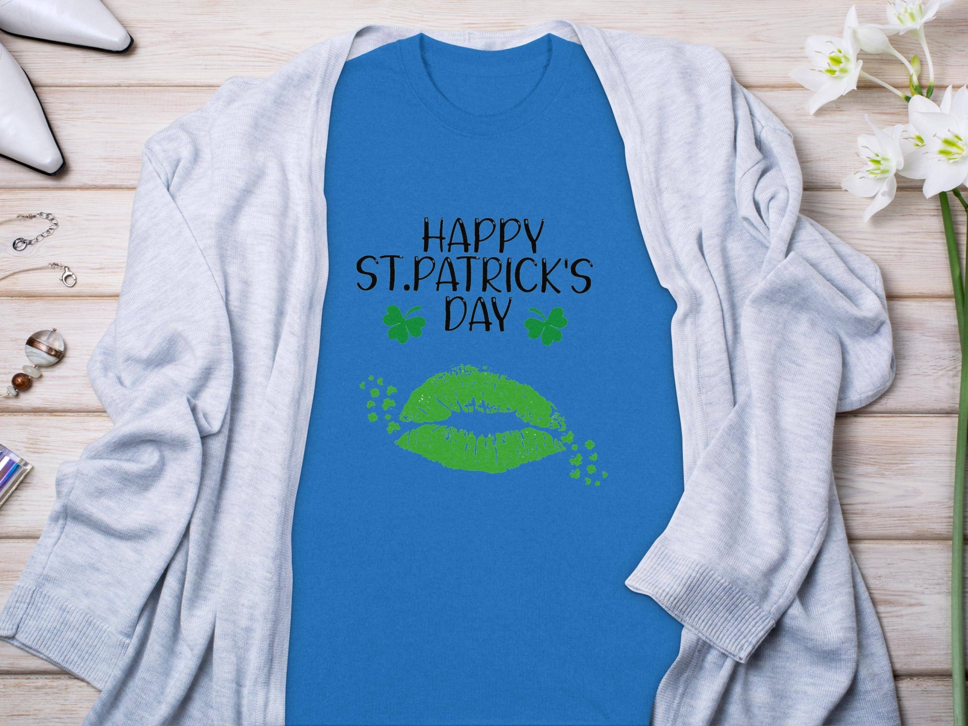 The Garment Graphics blue graphic T-shirt features green lips and clovers with Happy St. Patricks Day text. Its stylishly displayed on a wooden surface alongside a light gray cardigan, white flowers, and high-heeled shoes.