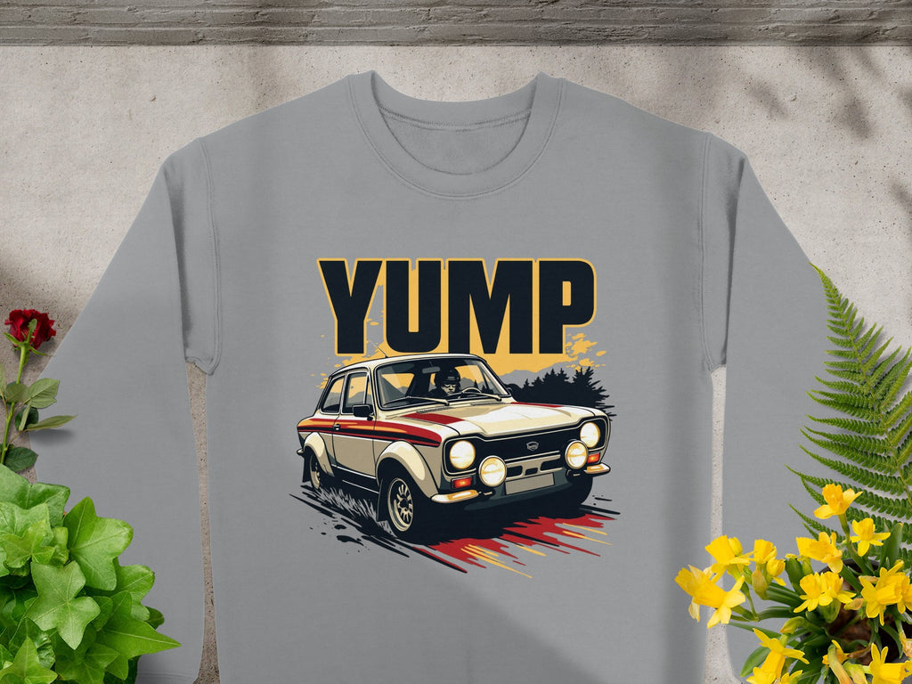 This Garment Graphics sweatshirt in gray showcases a bold YUMP design with a vintage rally car graphic. Laid flat on concrete, surrounded by green plants and yellow flowers, it exudes an effortlessly cool vibe ideal for car enthusiasts.
