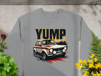 This Garment Graphics sweatshirt in gray showcases a bold YUMP design with a vintage rally car graphic. Laid flat on concrete, surrounded by green plants and yellow flowers, it exudes an effortlessly cool vibe ideal for car enthusiasts.
