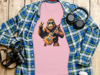 The Garment Graphics pink T-shirt showcases a guitar-playing, sunglasses-wearing Bigfoot making a peace sign, ideal for music lovers. Paired with a blue plaid shirt, camera, headphones, and coffee cup on wood; it brings an electric guitar flair to casual outfits.