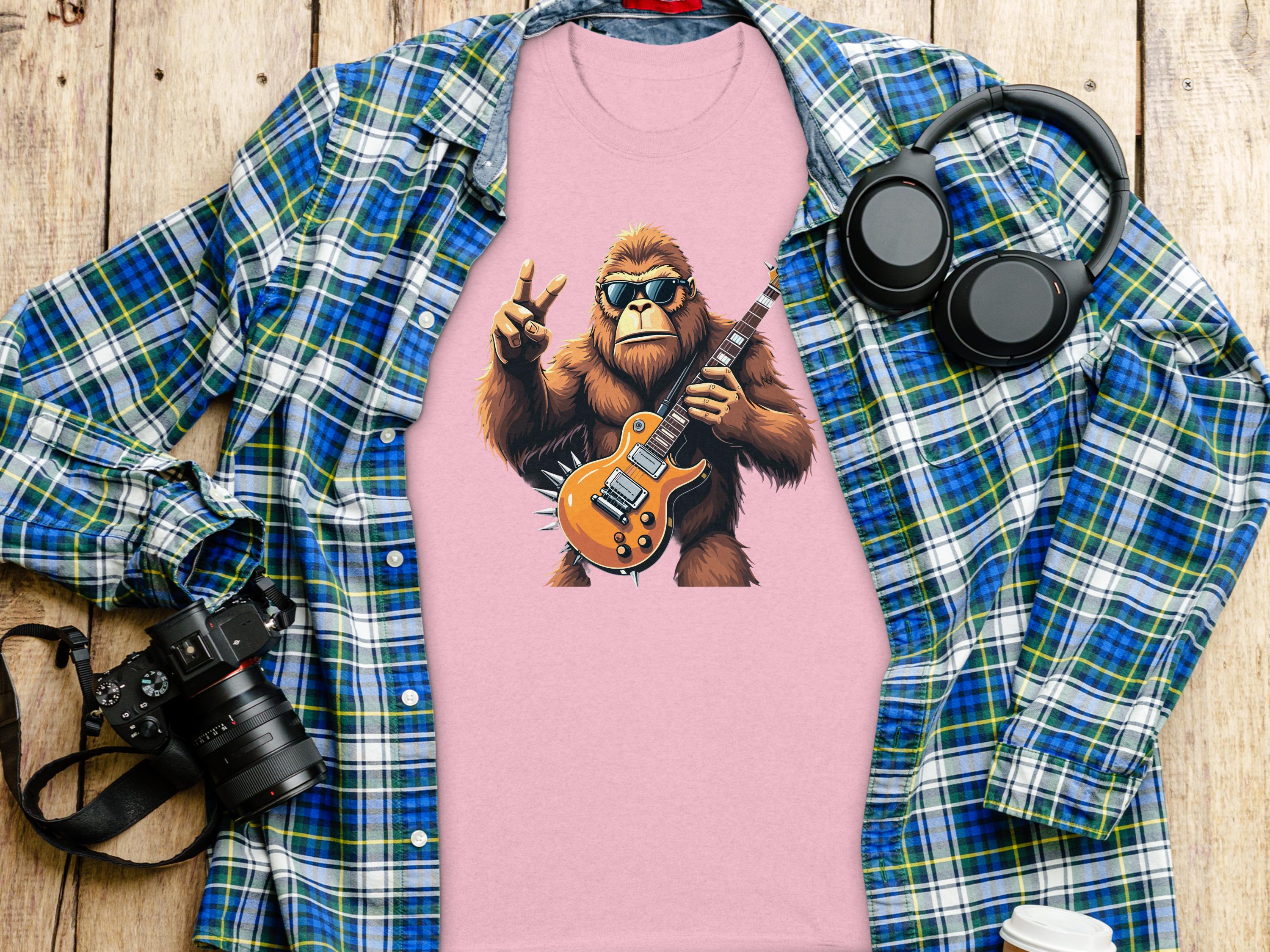 The Garment Graphics pink T-shirt showcases a guitar-playing, sunglasses-wearing Bigfoot making a peace sign, ideal for music lovers. Paired with a blue plaid shirt, camera, headphones, and coffee cup on wood; it brings an electric guitar flair to casual outfits.