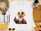 The Garment Graphics white cartoon eagle sweatshirt, in unisex sizing, shows a thumbs-up on a wooden surface. Nearby are a notepad with pen, scarf, sunglasses, coffee cup, and quilted jacket and purse—all coordinating in yellow and brown tones.