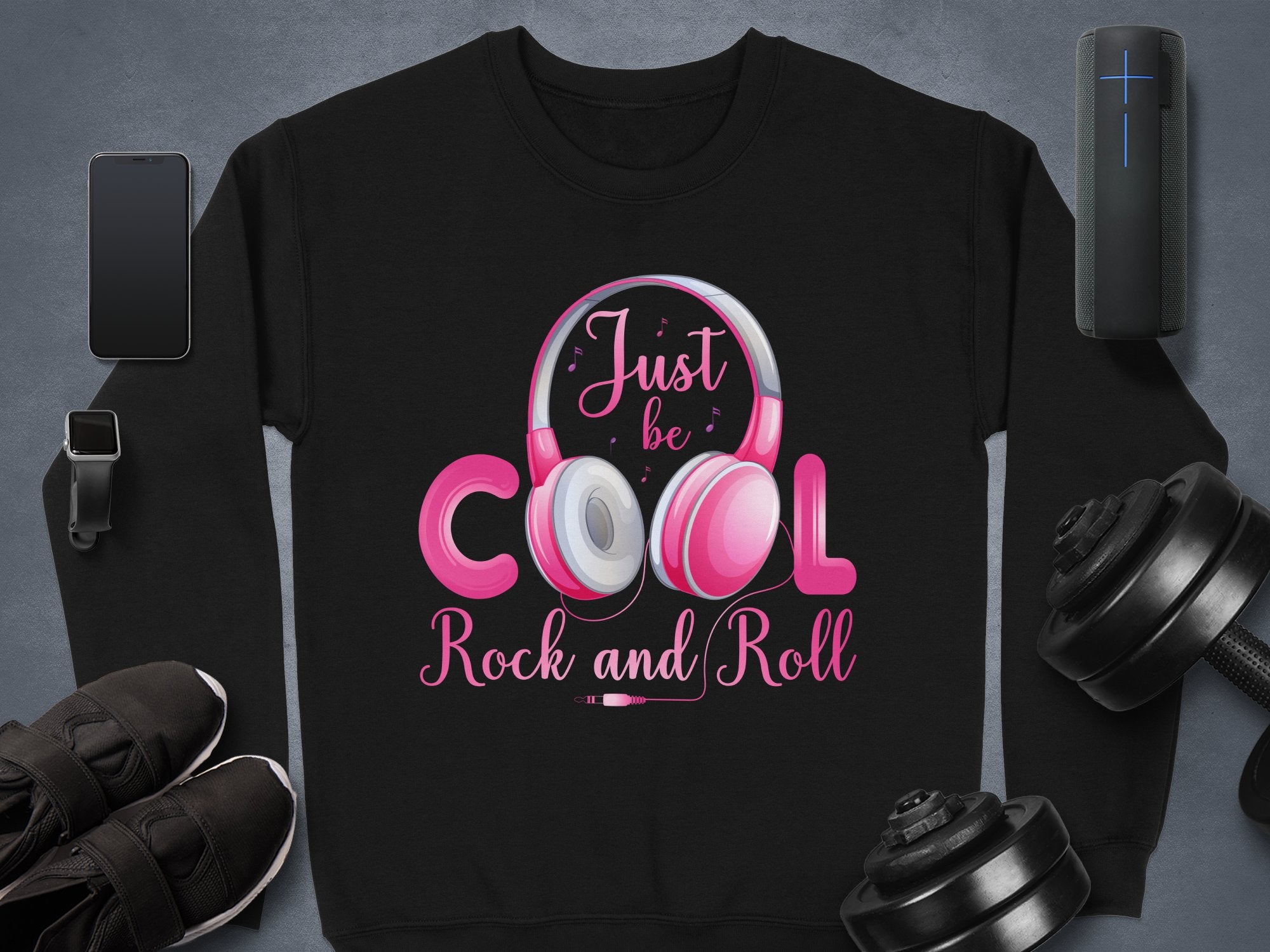 The Garment Graphics black sweatshirt, featuring Just be COOL Rock and Roll in pink and white with stylish headphones for music lovers, is perfect for a fashionable statement. Surrounded by a smartphone, smartwatch, speaker, black shoes, and dumbbells on a gray surface.