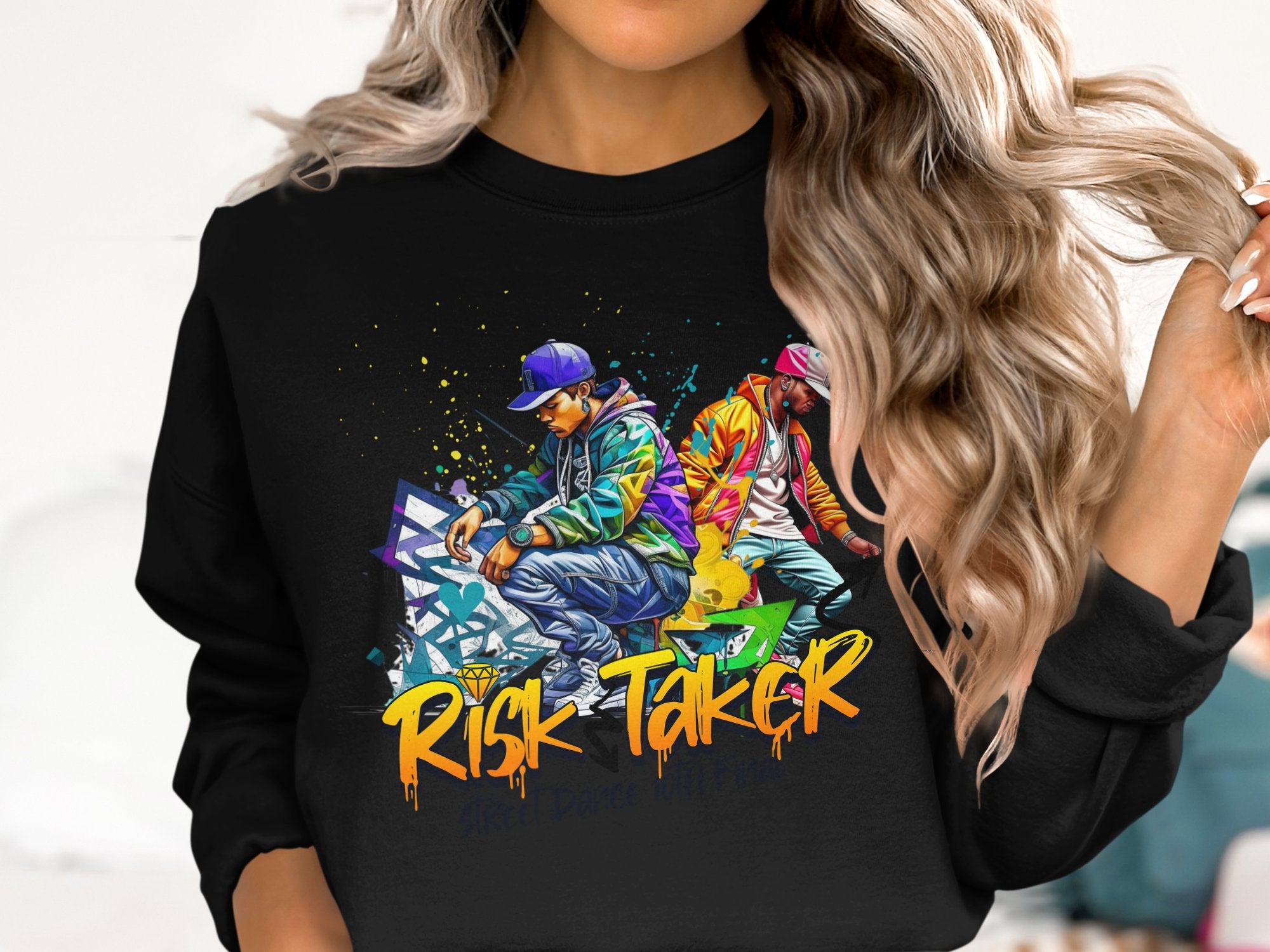 A person with long, wavy hair sports a black Garment Graphics sweatshirt featuring a vibrant graphic of two casually dressed figures. The bold graffiti-style text RISK TAKER Street Dance captures the dynamic spirit of street dance finals.