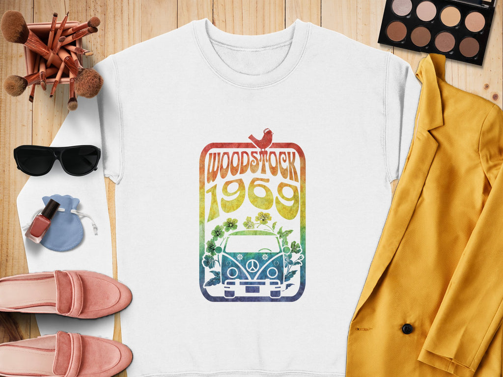 A flat lay features a white Garment Graphics sweatshirt with a retro Woodstock festival van and rainbow design. Surrounding it are pink loafers, sunglasses, cosmetics, nail polish, brushes, a blue pouch, and a mustard yellow blazer on a wooden surface.