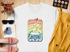 A flat lay features a white Garment Graphics sweatshirt with a retro Woodstock festival van and rainbow design. Surrounding it are pink loafers, sunglasses, cosmetics, nail polish, brushes, a blue pouch, and a mustard yellow blazer on a wooden surface.