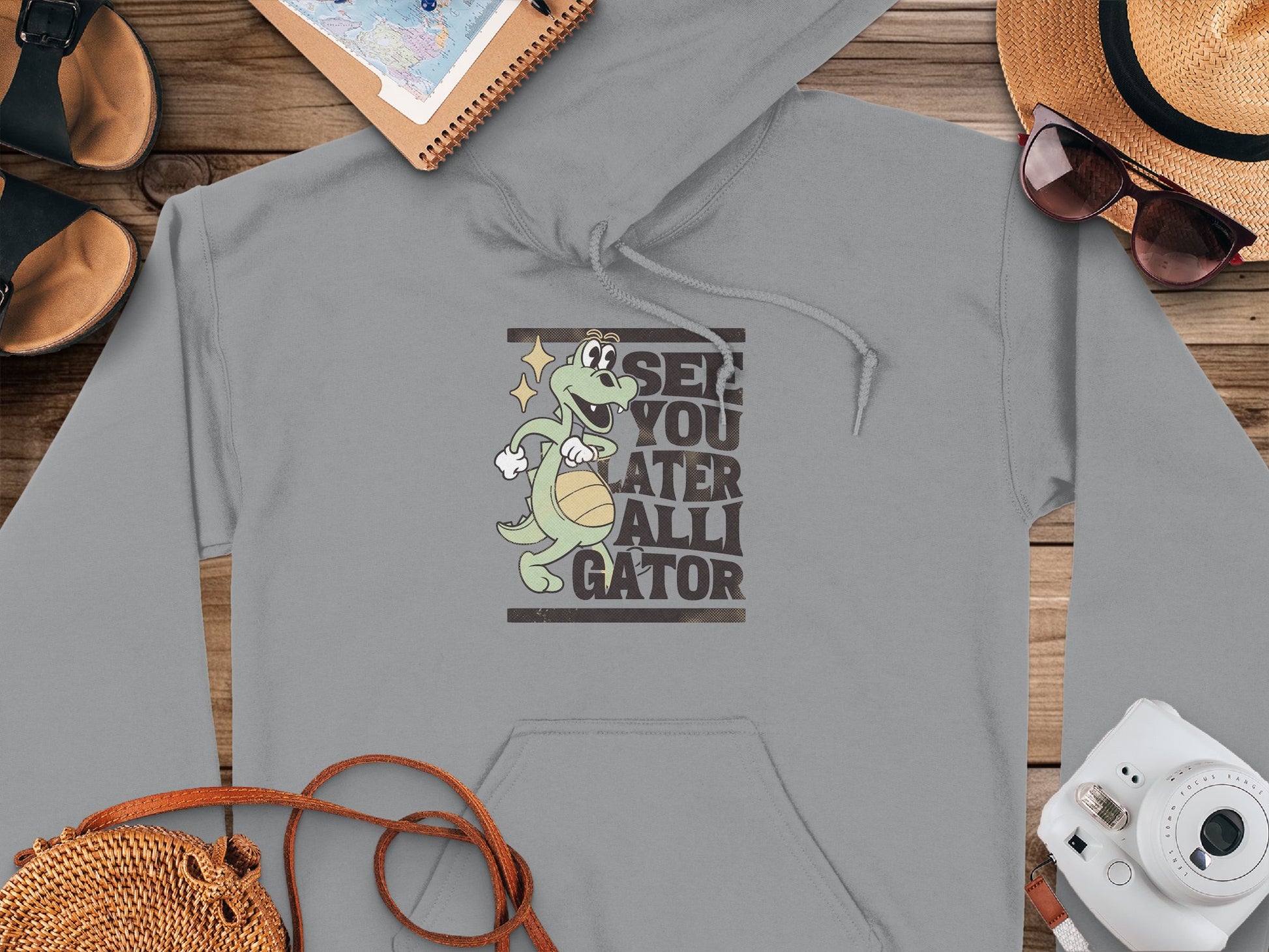 A classic-fit gray See You Later Alligator hoodie by Garment Graphics, showcasing a gator graphic with a basketball, rests on a wooden surface alongside travel essentials like a sun hat, sunglasses, map, sandals, camera, and woven bag—ideal for stylish adventures.