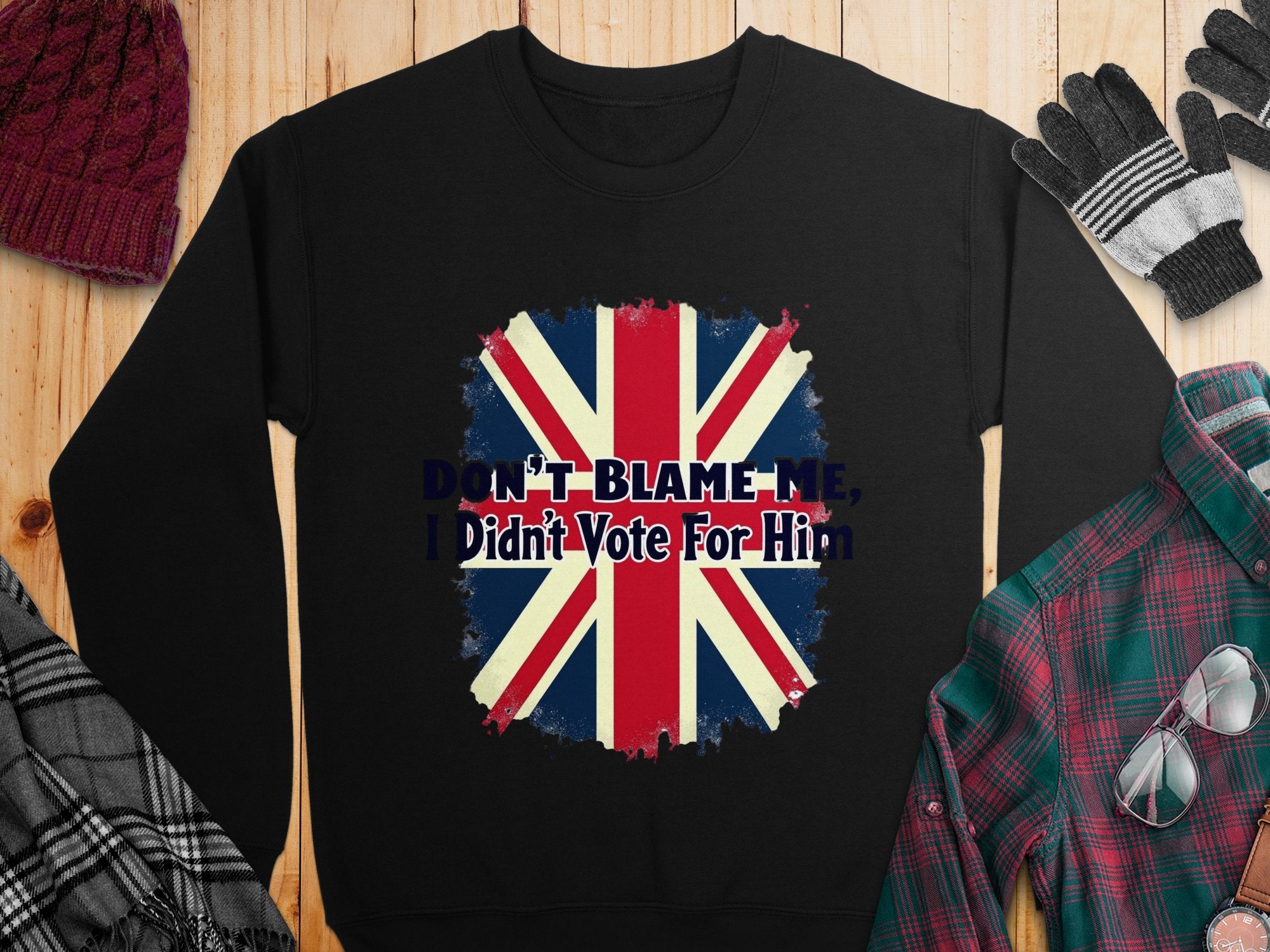 A black unisex Garment Graphics sweatshirt, featuring a bold Union Jack and the slogan DONT BLAME ME, I Didnt Vote For Him, is styled on wood with a scarf, hat, gloves, and plaid shirt for a trendy look.