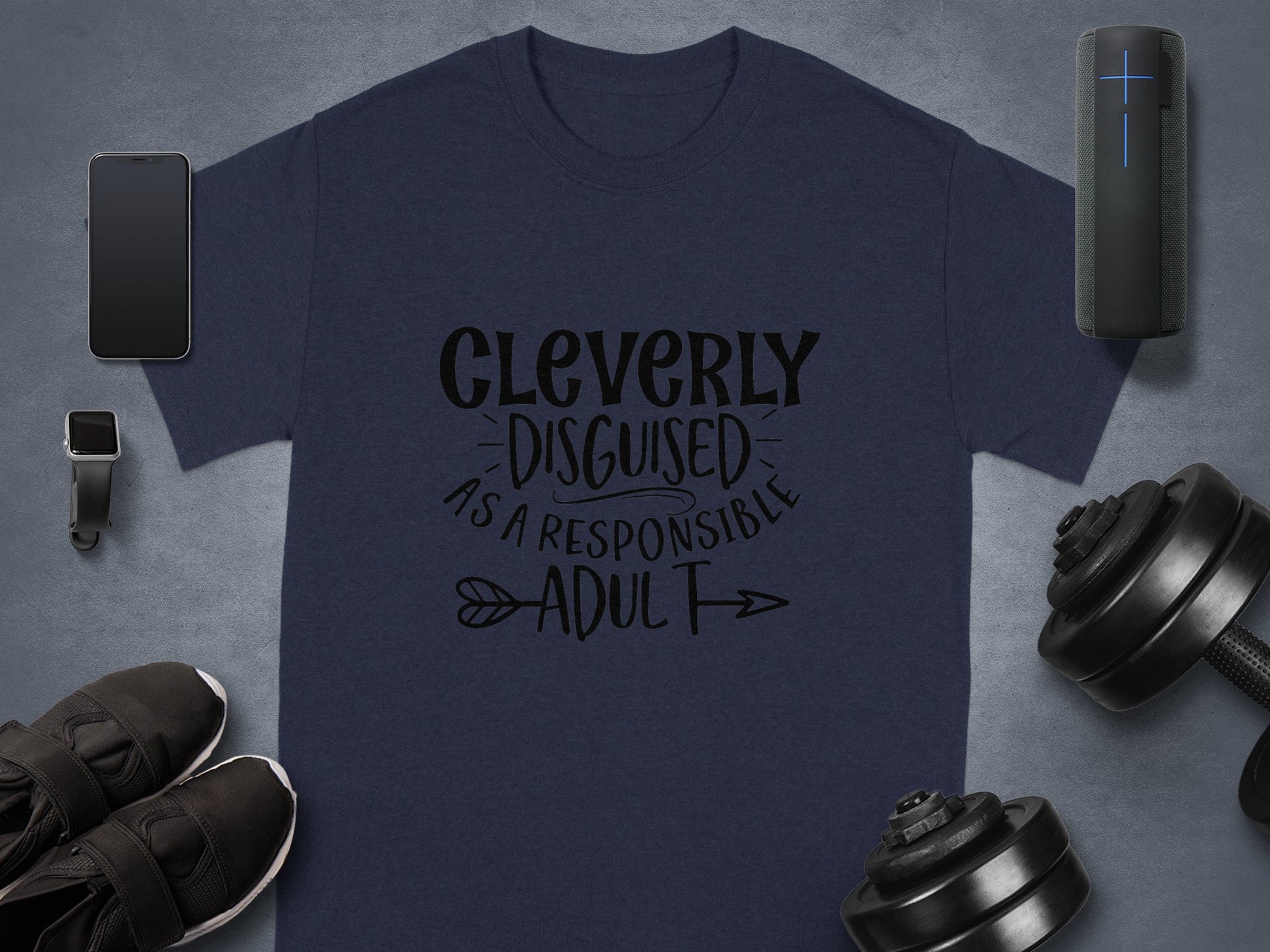 A Garment Graphics classic fit dark blue t-shirt with Cleverly Disguised as a Responsible Adult print is beautifully paired with a smartphone, smartwatch, earbuds, speaker, athletic shoes, and adjustable dumbbells—ideal for a casual outing on a gray background.