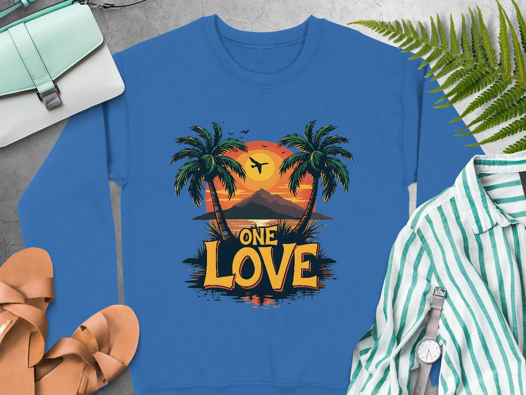 A Garment Graphics blue sweatshirt with palm trees, a sunset, and One Love Sunset text is pictured with sandals, a green striped shirt, a wristwatch, a fern leaf, and a blue handbag on a gray surface.