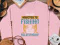 A pink unisex sweatshirt from Garment Graphics lies on a wooden surface, featuring the phrase: Im not addicted to fishing, were just in a very committed relationship. Beside it are a straw hat, sunglasses, a notepad, and a bag.