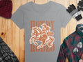 A gray Rodeo T-shirt by Garment Graphics features a cowgirl on horseback with Howdy Rodeo, Not a Beginner printed boldly. Laid on wood, its paired with a beanie, gloves, plaid shirt, and black-and-gray scarf—ideal for women’s sizing.