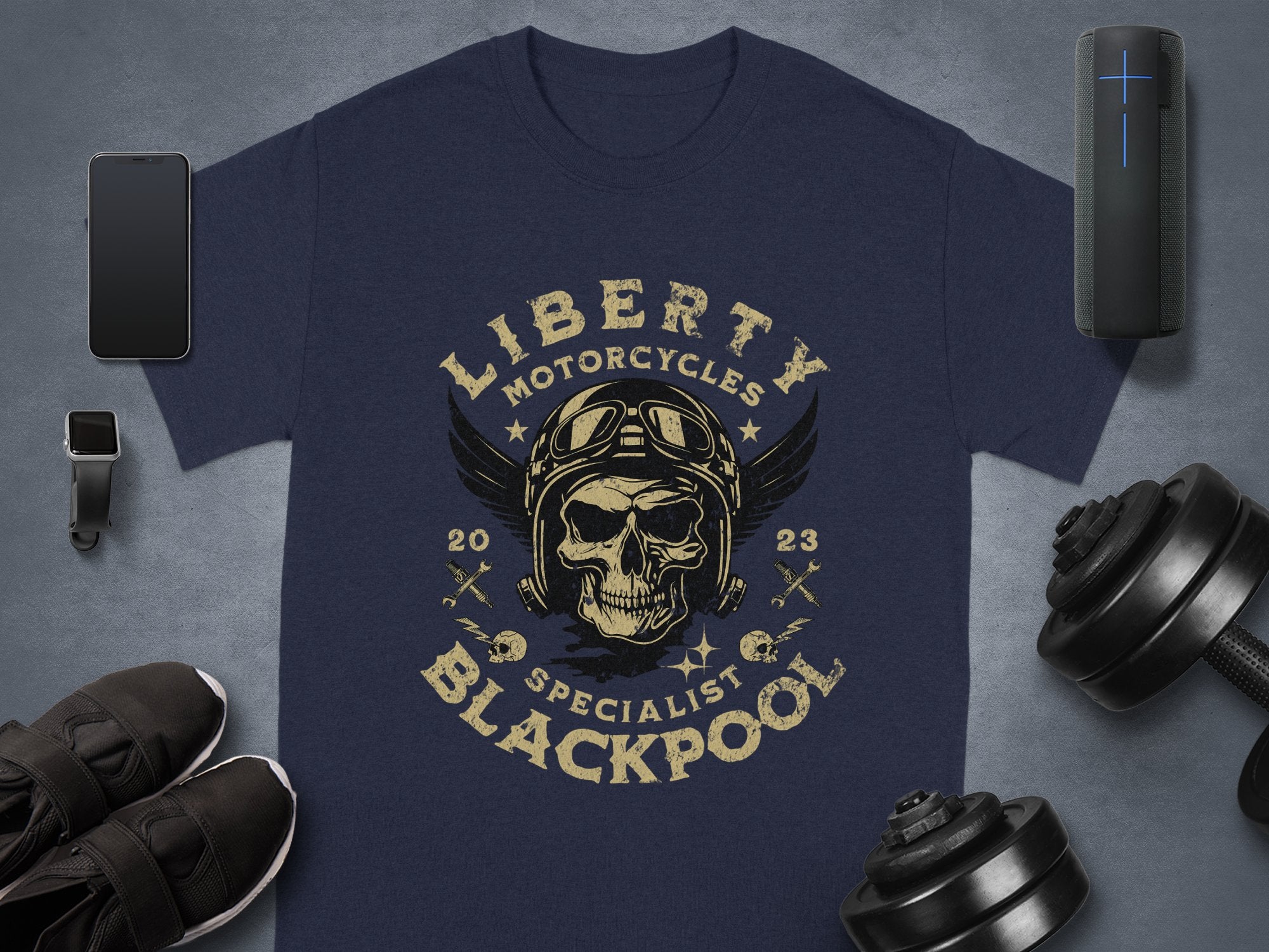 A Garment Graphics black T-shirt for motorcycle enthusiasts showcases a skull with goggles and wings, encircled by Liberty Motorcycles Specialist Blackpool 2023. Its laid out with a phone, smartwatch, sneakers, headphones, and dumbbells nearby.