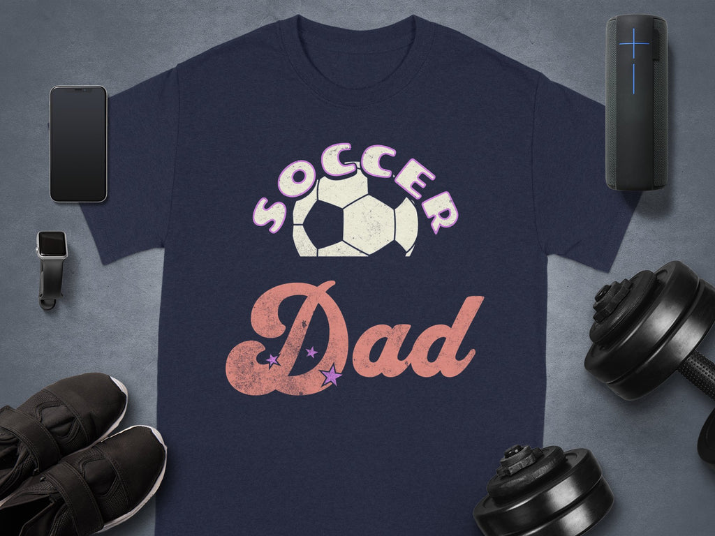 The Garment Graphics navy blue Soccer Dad T-shirt features a bold soccer ball graphic on a dark background, accompanied by a smartphone, smartwatch, wireless speaker, sleek black sneakers, and adjustable dumbbells.