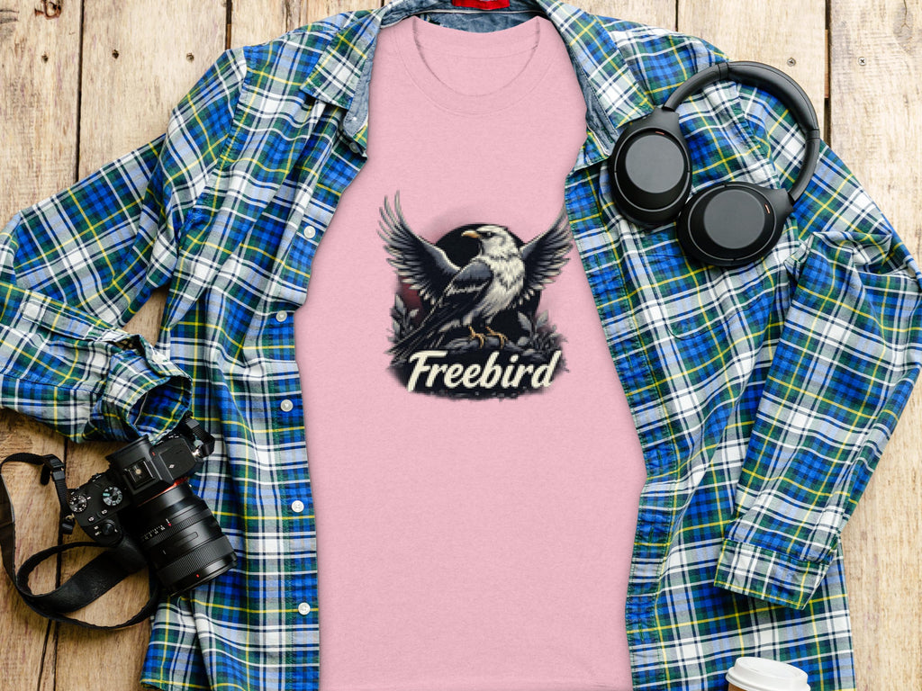 A pink Freebird eagle T-shirt by Garment Graphics, made of 100% cotton with a bold bird graphic, lies on a wooden surface. Nearby are a blue plaid shirt, camera, headphones, and white cup.