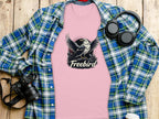 A pink Freebird eagle T-shirt by Garment Graphics, made of 100% cotton with a bold bird graphic, lies on a wooden surface. Nearby are a blue plaid shirt, camera, headphones, and white cup.