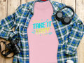 A Take It Easy T-Shirts pink top from Garment Graphics, with vibrant graphics, sits under a blue/green plaid shirt. Right: headphones; left: camera. A coffee cup and wooden background complete this chic design.
