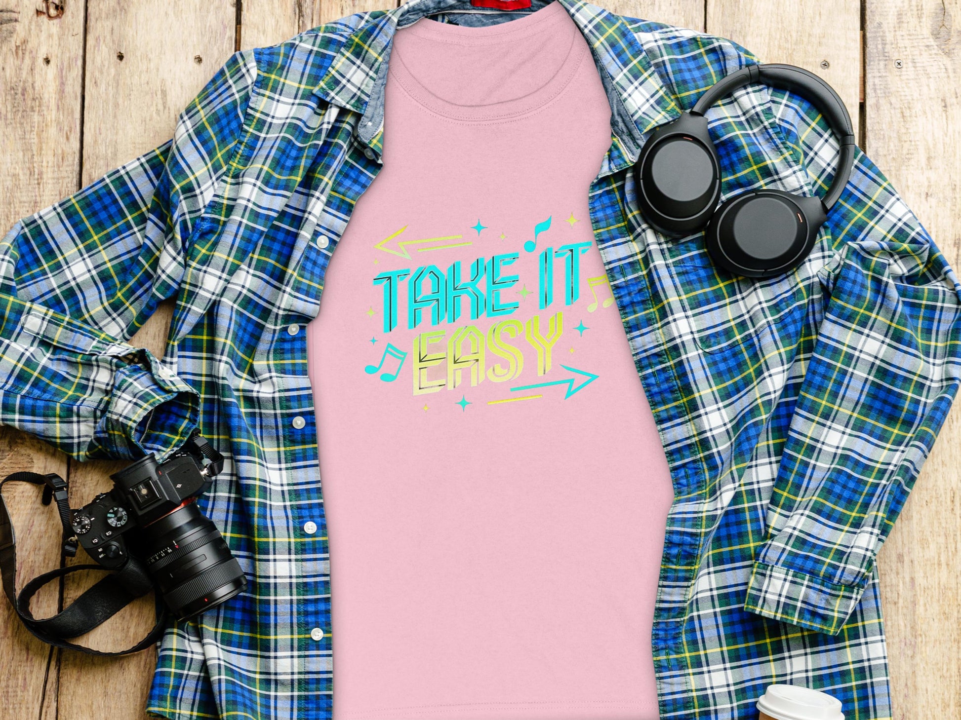 A Take It Easy T-Shirts pink top from Garment Graphics, with vibrant graphics, sits under a blue/green plaid shirt. Right: headphones; left: camera. A coffee cup and wooden background complete this chic design.
