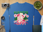 This loose-fit blue sweatshirt from Garment Graphics Hands Off Wild Side collection features a cartoon woman in a hat with a gun, bold HANDS OFF! text, and floral accents. Displayed with a camera, lens, cucumbers, and notebook on wood.