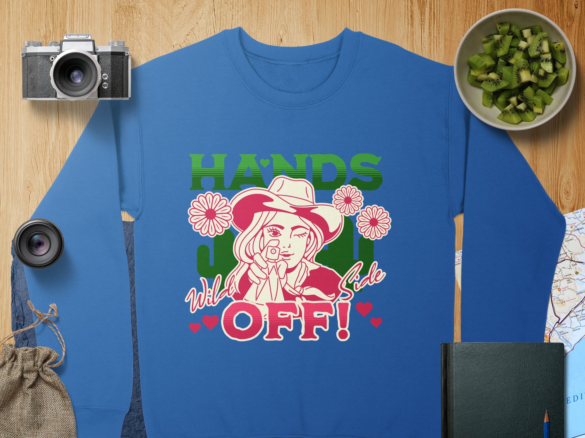 This loose-fit blue sweatshirt from Garment Graphics Hands Off Wild Side collection features a cartoon woman in a hat with a gun, bold HANDS OFF! text, and floral accents. Displayed with a camera, lens, cucumbers, and notebook on wood.