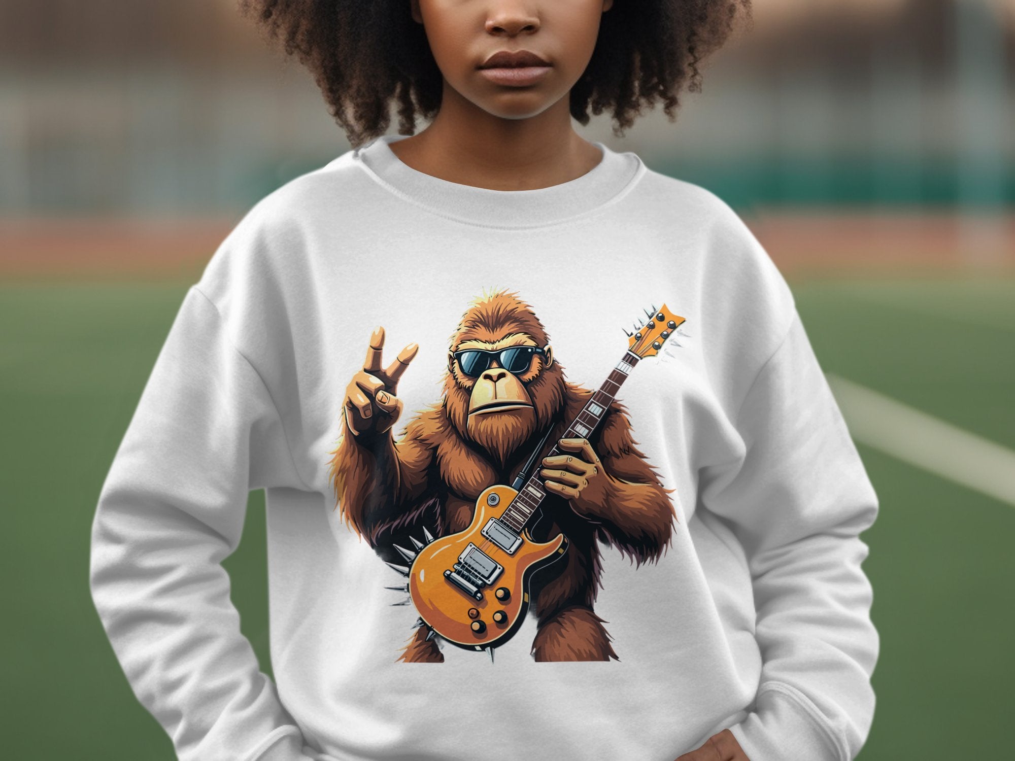 A person is wearing a Garment Graphics sweatshirt with a gorilla holding an electric guitar and flashing a peace sign, complete with sunglasses, set against an outdoor sports field.