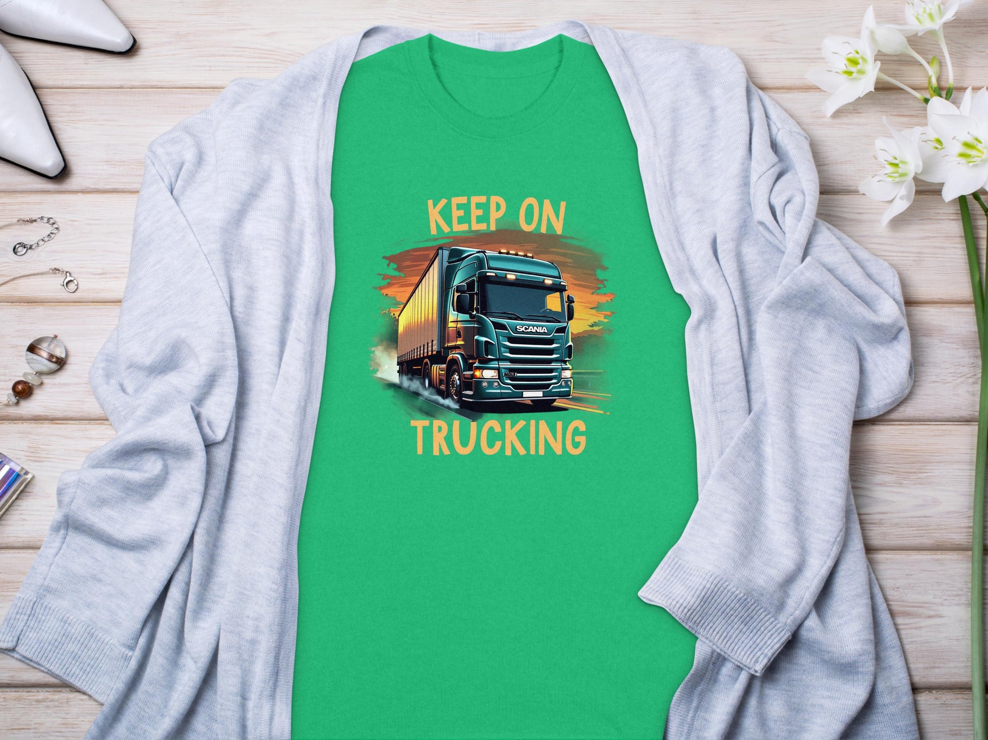 The Garment Graphics green graphic T-shirt for truck enthusiasts features a large truck illustration with Keep On Trucking text. It is displayed on wood, paired with a light gray cardigan, white flowers, high heels, and jewelry.