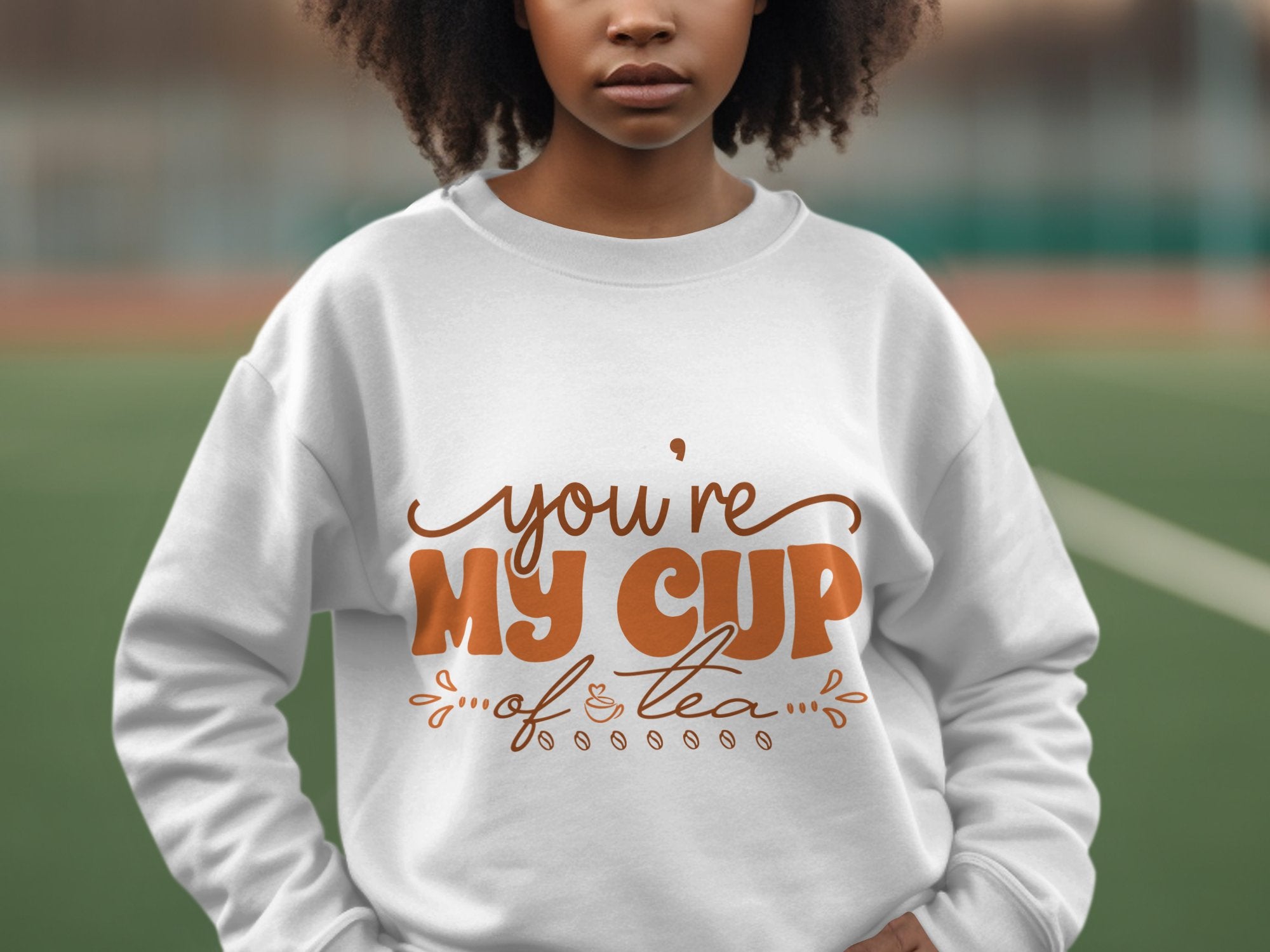 A person stands outdoors in a cozy Garment Graphics sweatshirt, featuring the phrase Youre my cup of tea in brown cursive. The unisex-sized apparel is set against a blurred background with hints of a tennis court and fencing in the distance.