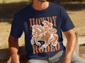 A person is sitting on a wooden bench, proudly wearing a navy blue Garment Graphics T-shirt featuring a cowgirl riding a horse with the bold brown words Howdy Rodeo and Not a Beginner.