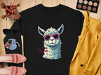 A flat lay features Garment Graphics No Prob-Llama sweatshirt, boasting a cartoon llama in sunglasses. Surrounding it are a yellow blazer, makeup brushes, eyeshadow palette, blue pouch, sunglasses, lipstick, and pink shoes—ideal for fans of unisex sizing and quirky fashion!.