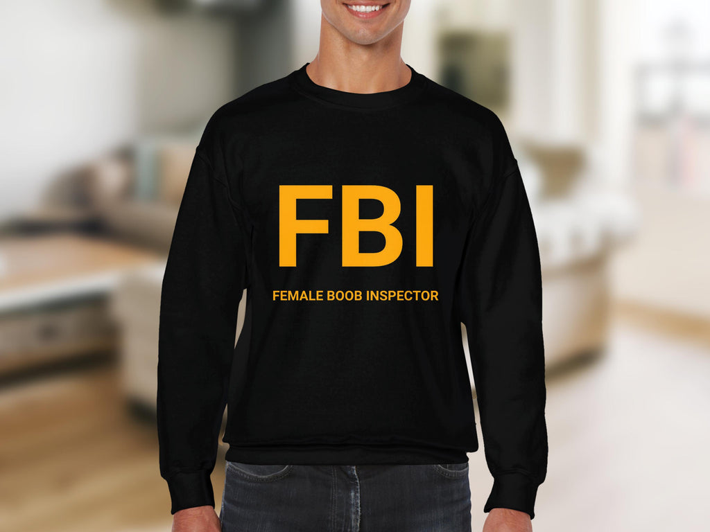 Wearing a Garment Graphics black sweatshirt with bold yellow FBI and Female Boob Inspector text, a man confidently stands in a room with a blurred background. The unisex sizing ensures a comfortable fit.