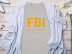 A gray Garment Graphics T-shirt features a fun design with bold orange FBI letters and Female Boob Inspector below. Its paired with a light blue cardigan, white shoes, flowers, and small accessories on a wooden surface.