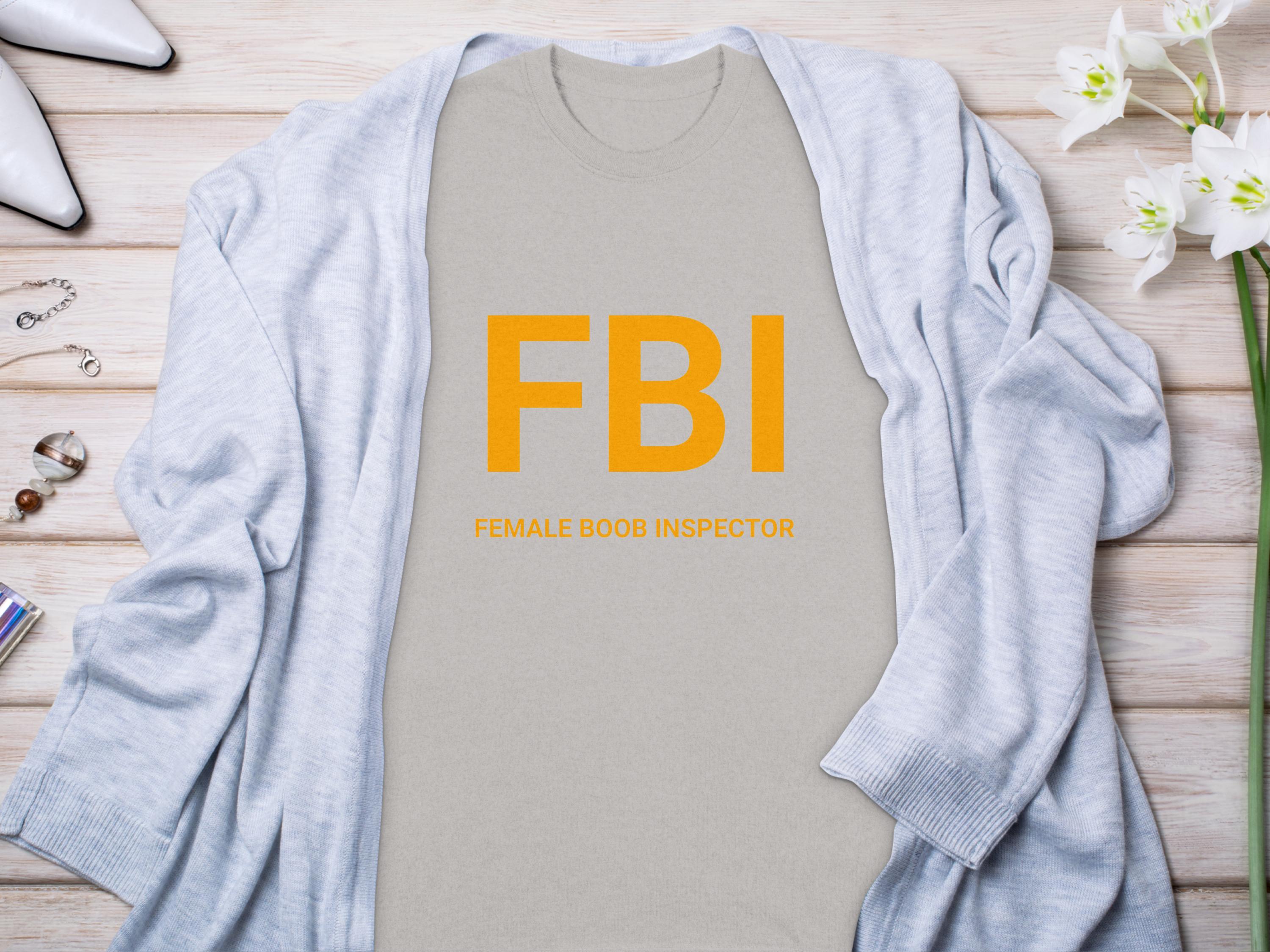 A gray Garment Graphics T-shirt features a fun design with bold orange FBI letters and Female Boob Inspector below. Its paired with a light blue cardigan, white shoes, flowers, and small accessories on a wooden surface.