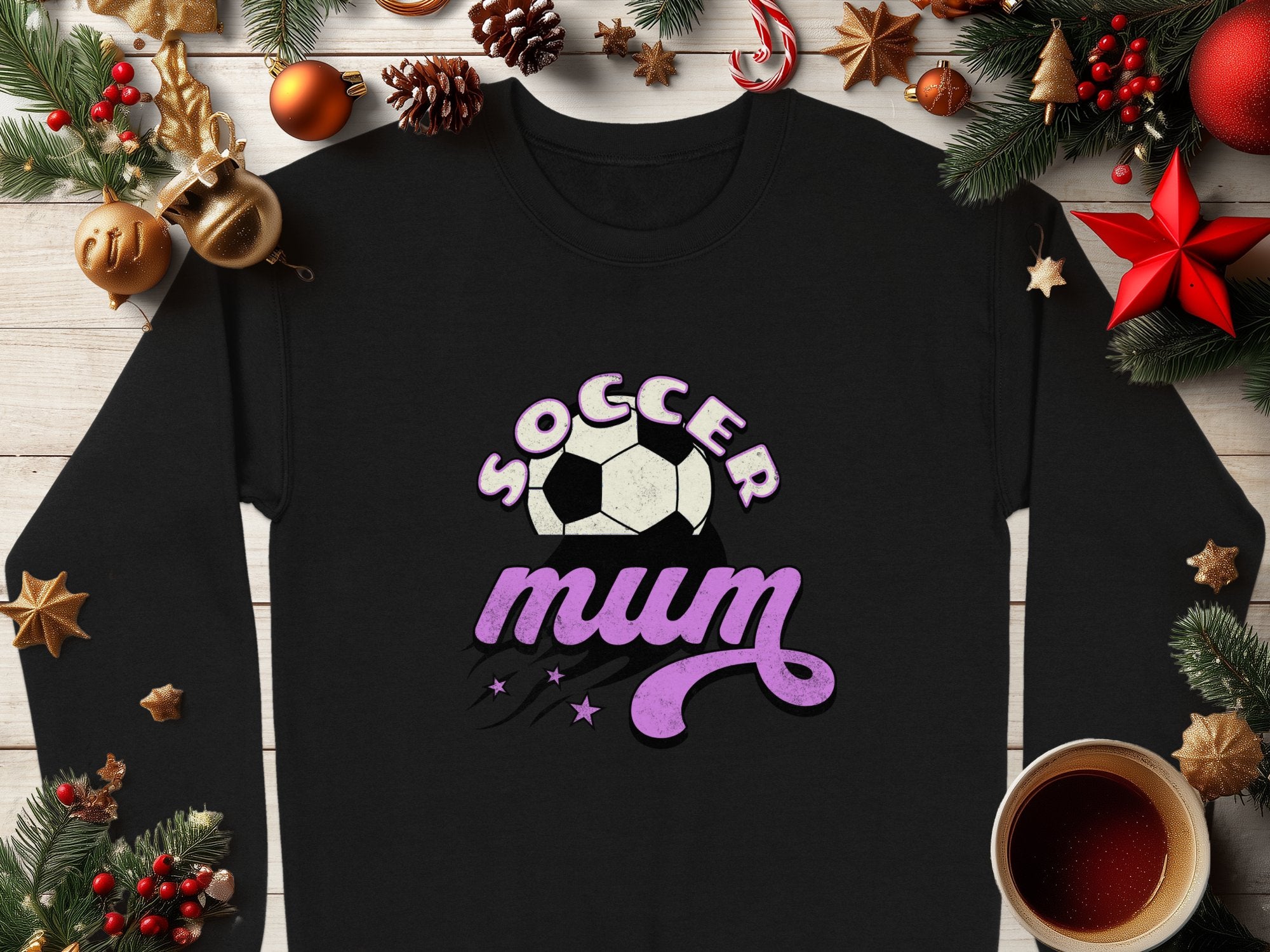A Garment Graphics black sweatshirt, featuring Soccer Mum in white and purple vintage style with a soccer ball graphic, is set among festive decorations like pine cones, ornaments, a red star, and a steaming coffee cup on wood.
