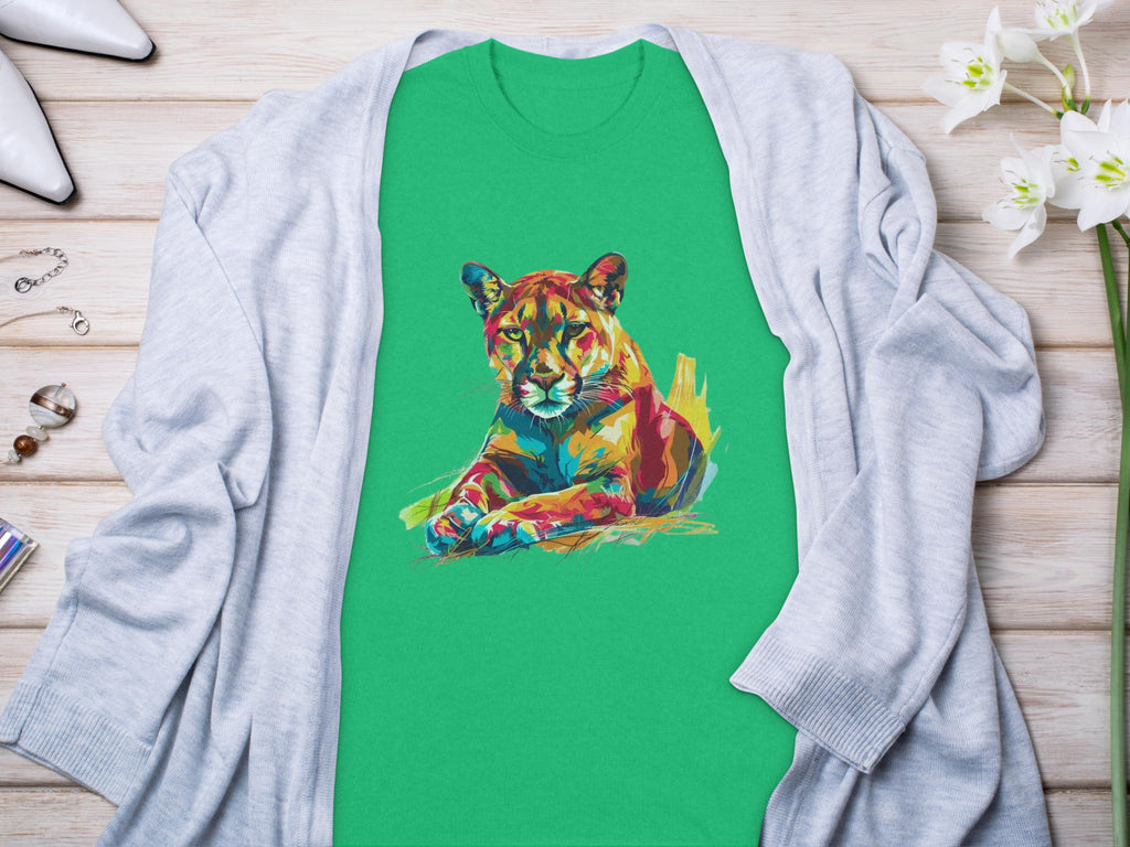 A vibrant green T-shirt from Garment Graphics showcases an abstract lioness design. Its styled laid flat on light wood, accented with a gray cardigan, white heels, jewelry, and white flowers.