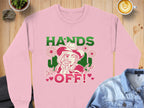 The Wild Side Sweatshirt by Garment Graphics, showcasing bold graphics of a person in a hat with flowers and HANDS OFF! text, is displayed on wood. Nearby are a latte, denim shirt, small plant, and rock. Its available in unisex sizing for adventurous souls.