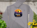 The Garment Graphics gray hoodie displays a vivid sunset mountain design with a backpacker silhouetted against the sun, featuring the phrase Retired 2025: Not My Problem Anymore, and is set against concrete with lush greenery surrounding it.