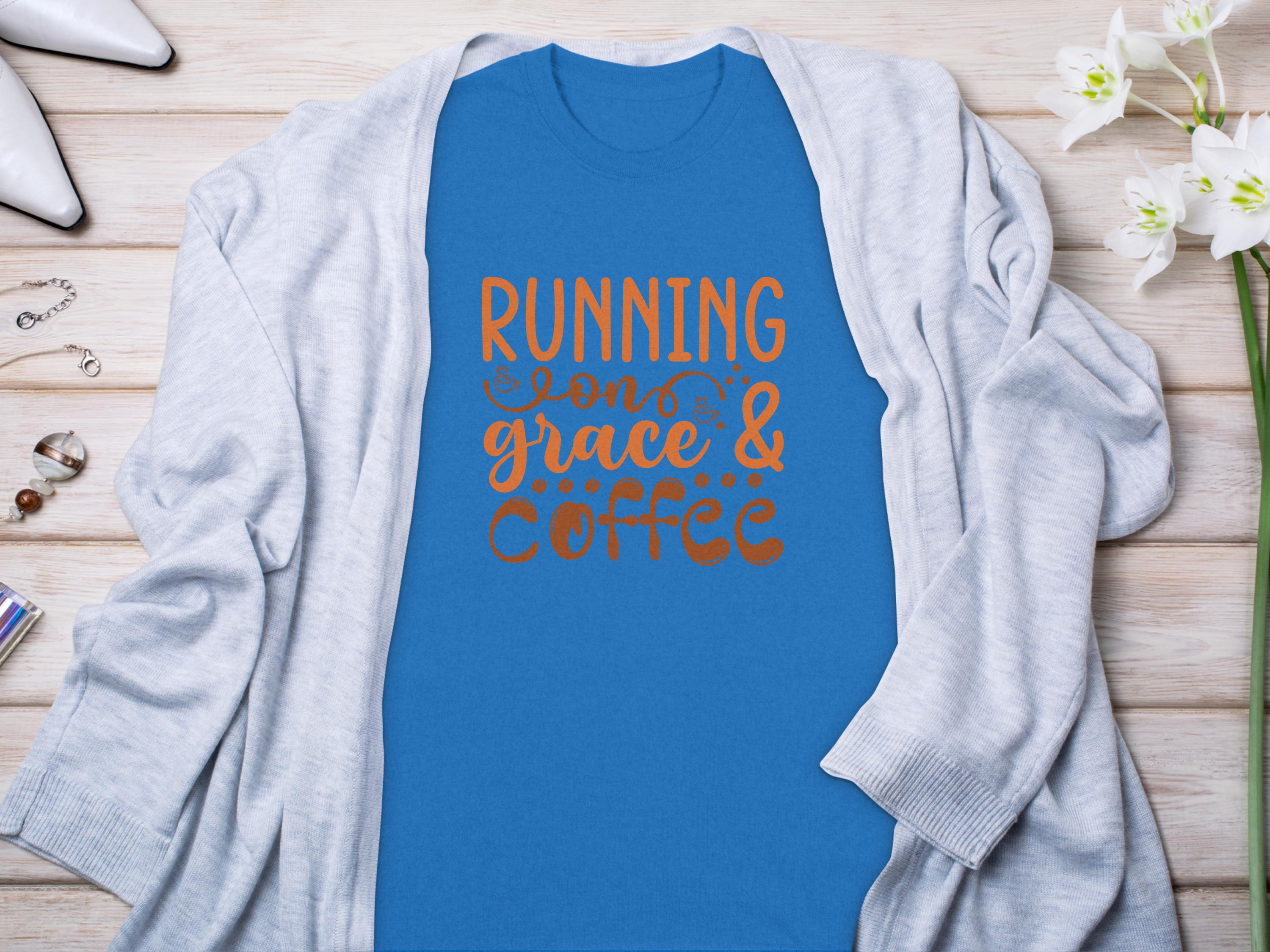 A blue Garment Graphics T-shirt, featuring the quote RUNNING on grace & coffee in orange, is styled under a light gray cardigan on wood, with white flowers and jewelry nearby.