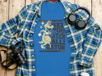 A blue t-shirt from Garment Graphics showcases a cartoon alligator with a basketball and the text See You Later Alligator. Its displayed on a plaid shirt with essentials: camera, headphones, and white cup on a wooden surface.