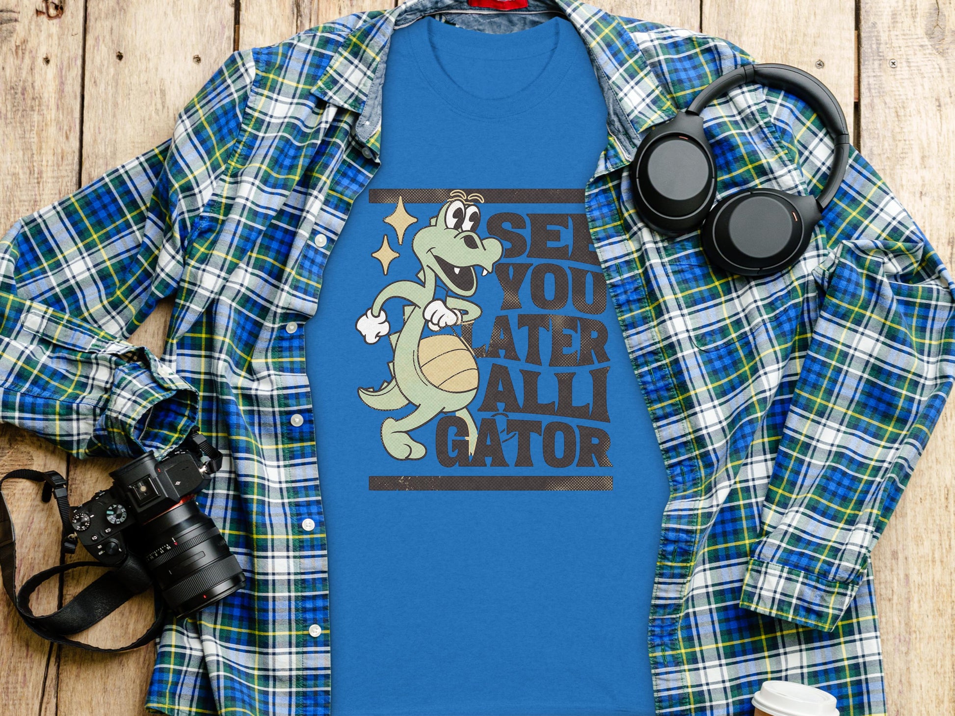A blue t-shirt from Garment Graphics showcases a cartoon alligator with a basketball and the text See You Later Alligator. Its displayed on a plaid shirt with essentials: camera, headphones, and white cup on a wooden surface.